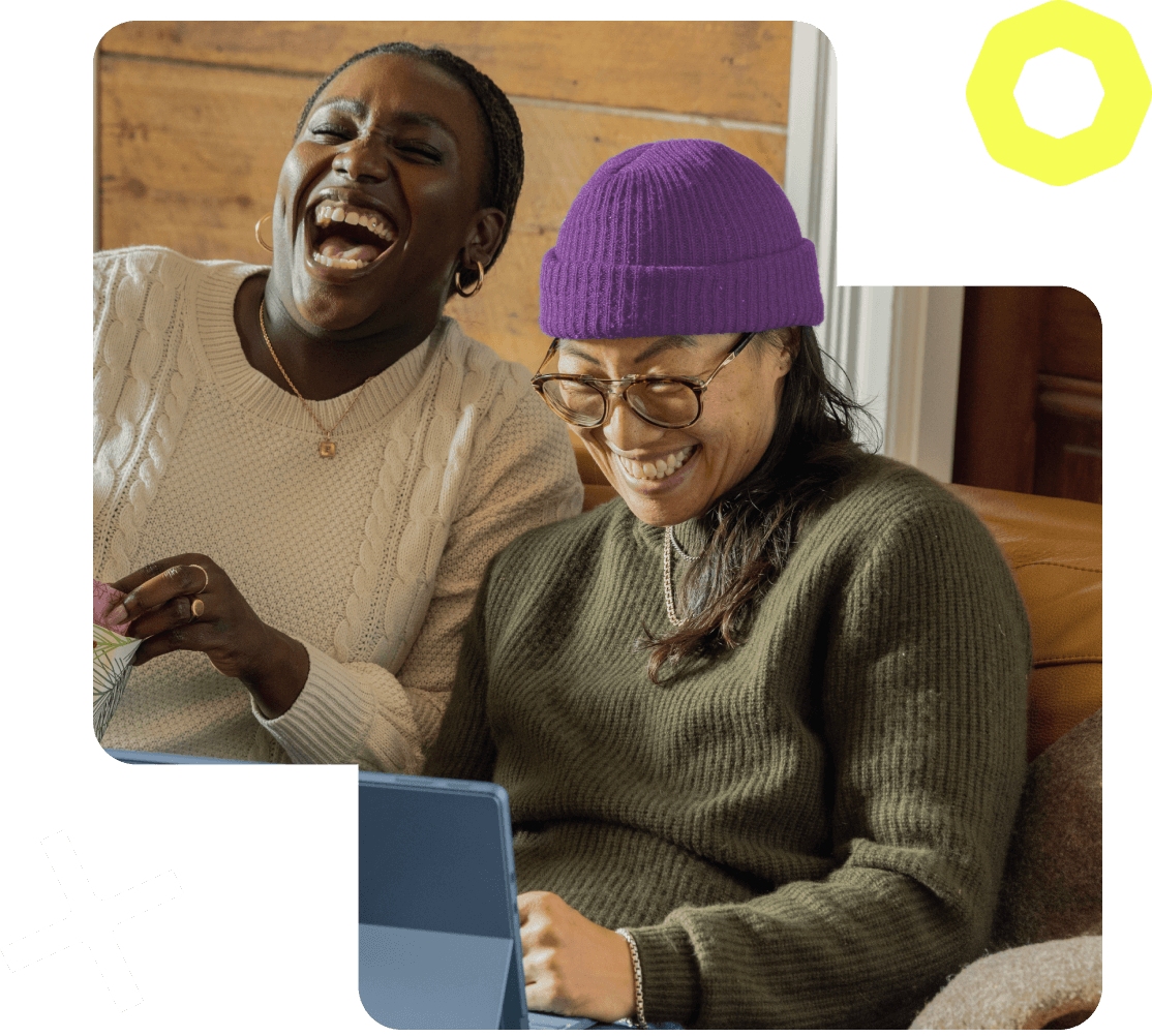 Image of two people sitting together on a sofa, laughing while working on a laptop. 